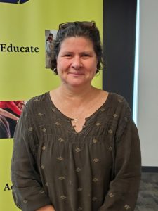 Judy McGuigan, fair/white skin, glasses on hair, aging brown hair with white and darker streaks, green shirt with decorations and wearing a necklace and earings in front of a yellow banner