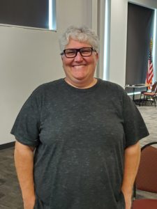fair/white woman with black framed glasses and white hair. Wearing a greyish shirt.
