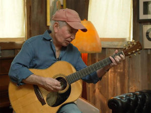 Singer-Songwriter Paul Simon playing a guitar with a faded red cap and blue shirt.  Image source from CBS News.