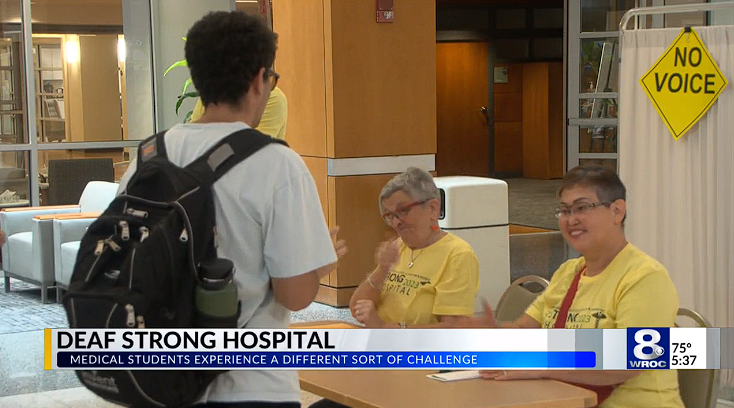 Yellow sign in background says 'No Voice' as 2 people in yellow shirts check in a patient as part of the program.
