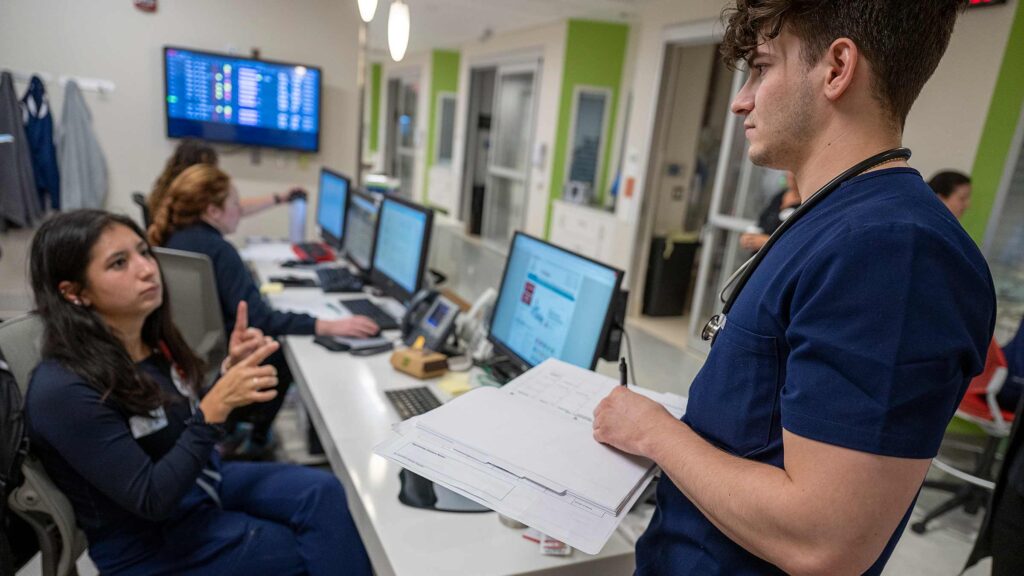 Pushing the limits of what’s possible, Casey King sets an example for other deaf people interested in medical careers.  

Casey, seated communicates with a male nurse standing up on the right.  Computer screens are seen between them.  