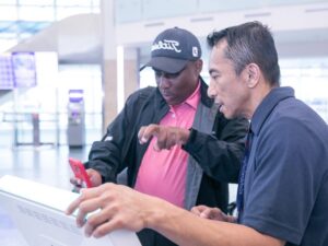 Left - black man wearing hat and using a red phone with ASL to communicate with airport worker on right.