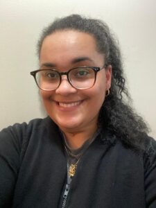 Alanna Smith with black curly hair wearing a black jacket and a necklace with a tan/beige colored background