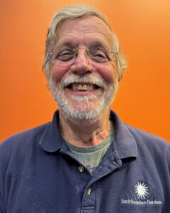 Dave with white hair and beard. Wearing transparent glasses and blue Smithsonian Garden shirt with orange background