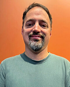 Feta with brown hair and a mixed colored goatee of brown and white. Wearing green shirt with orange background