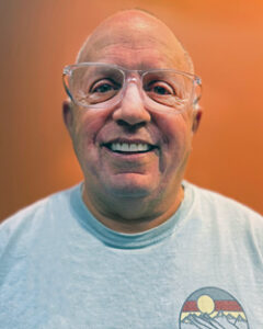 Fred Hartman with extremely short white hair smiling with whitish glasses and wearing a blue shirt with orange background.