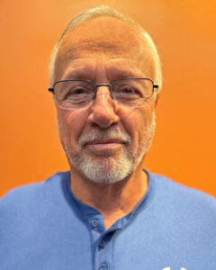 Jay Innes with white hair and goatee wearing glasses and blue shirt with orange background.