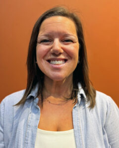 Kelli with brown hair smiling and necklace. Wearing a blue shirt over a white shirt with orange background