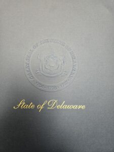 Delaware State Governor's seal is shown - center shows 2 people side by side with a cow in the center of a plaque which is similar or same as the state flag or seal
